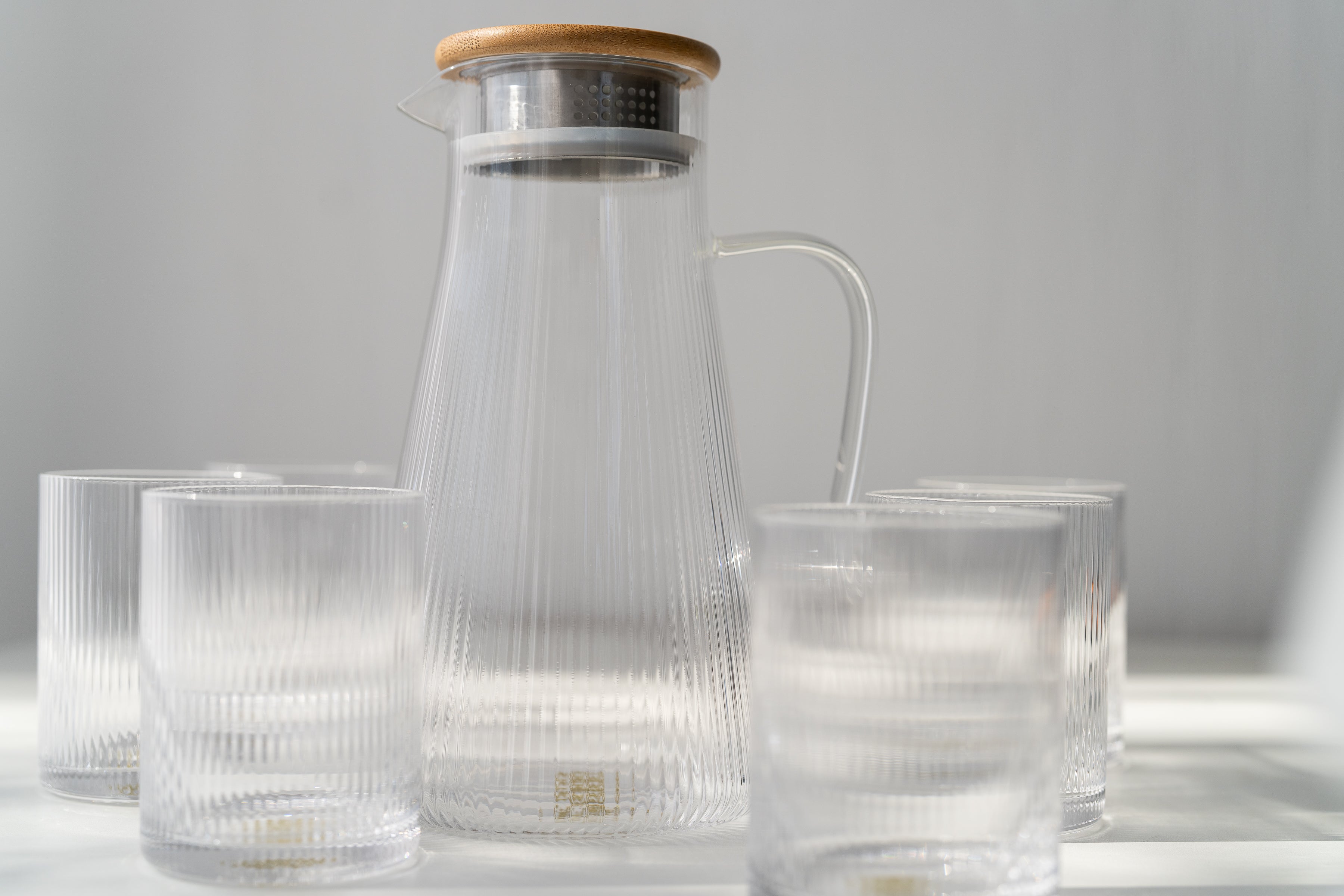 Clear glass pitcher with wooden lid and matching glasses on a light gray background