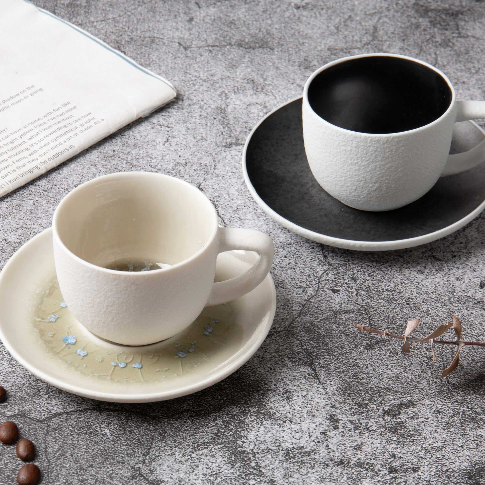 Two ceramic cups on saucers on a textured surface with coffee beans and a newspaper.