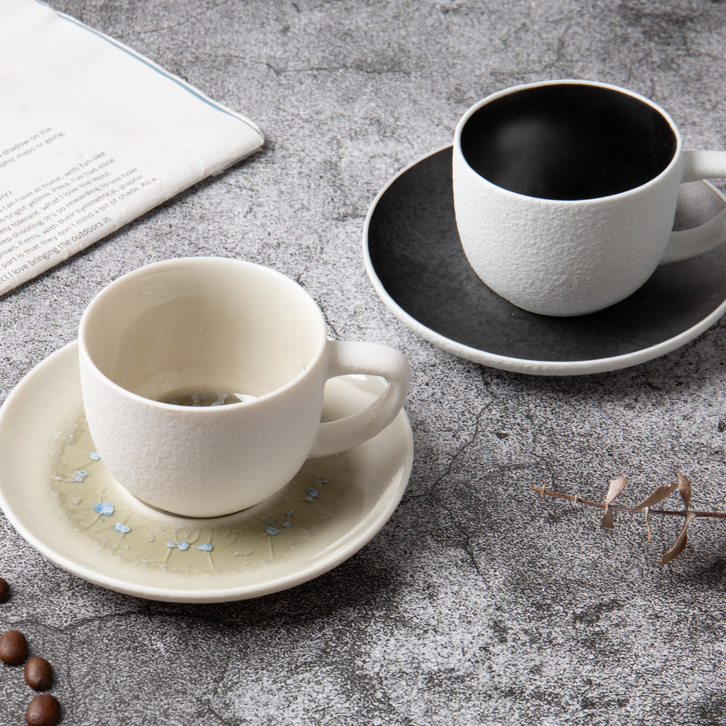 Two ceramic cups on saucers on a textured surface with coffee beans and a newspaper.