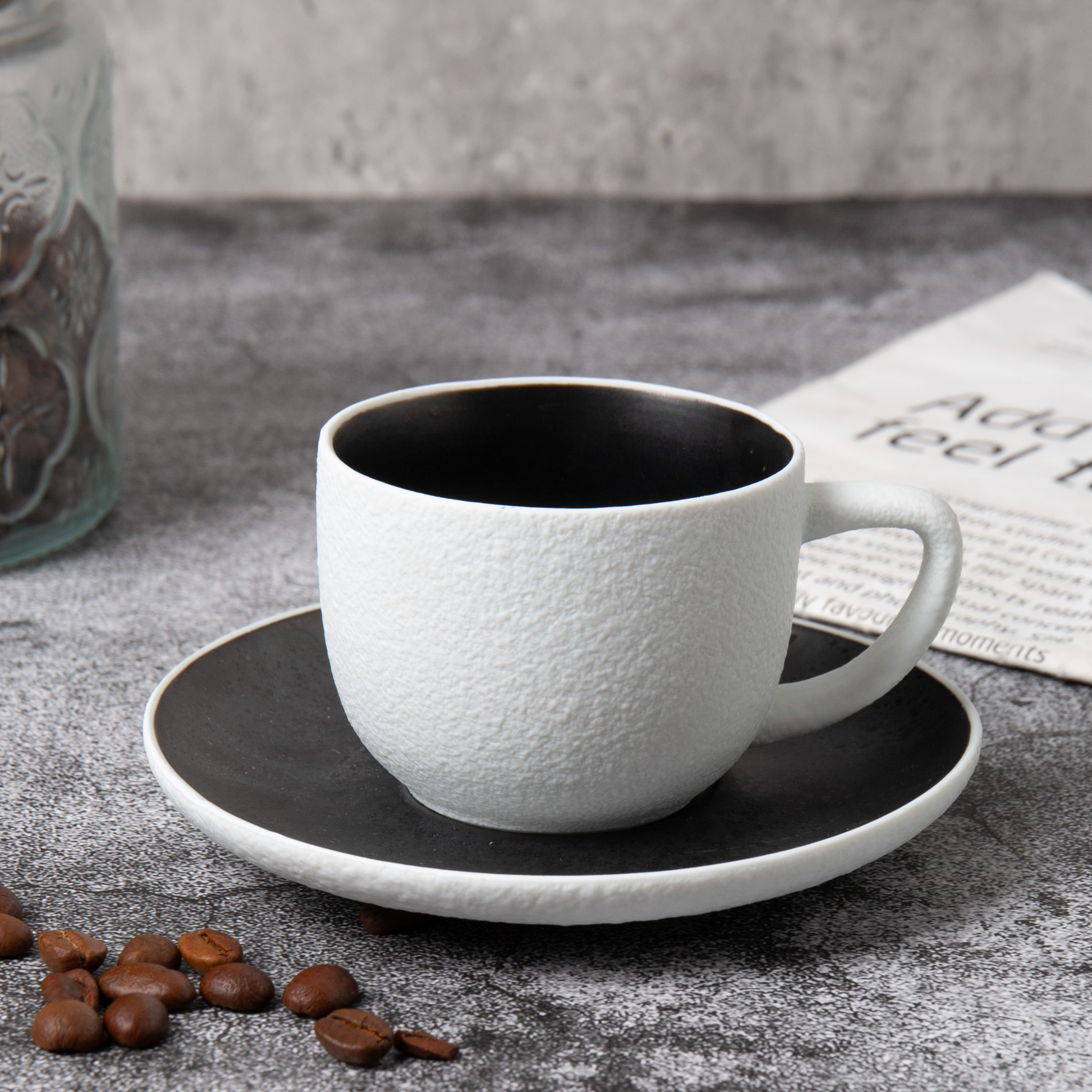 White textured coffee cup and saucer on a gray surface with coffee beans and a jar in the background.